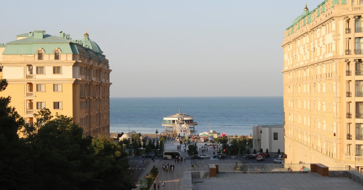 Open water is in the distance. A crowd on the beach can be seen between two tall buildings.