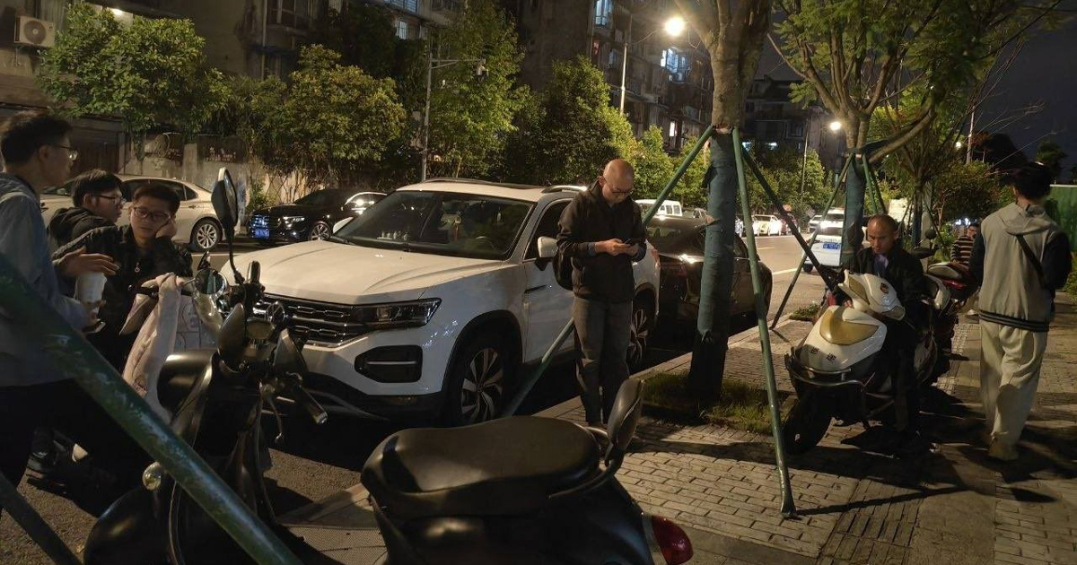 Nighttime urban street photograph showing pedestrians, parked scooters, trees, and a white SUV. Streetlights illuminate people gathered near the vehicles.
