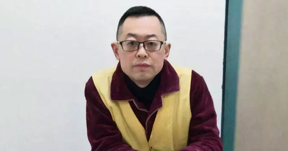 Photograph of Pastor Wang Yi seated indoors, wearing a burgundy shirt and yellow safety vest. A plain light wall and teal doorframe form the background.