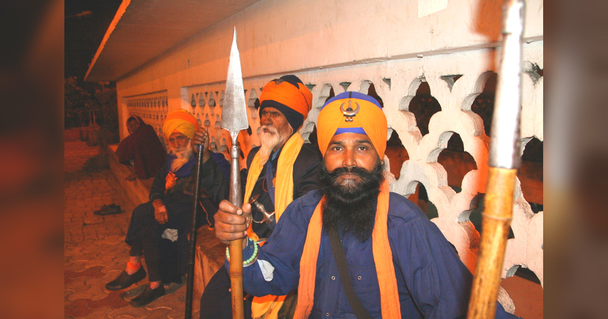Men dressed in orange and blue turbans and robes, holding spears.