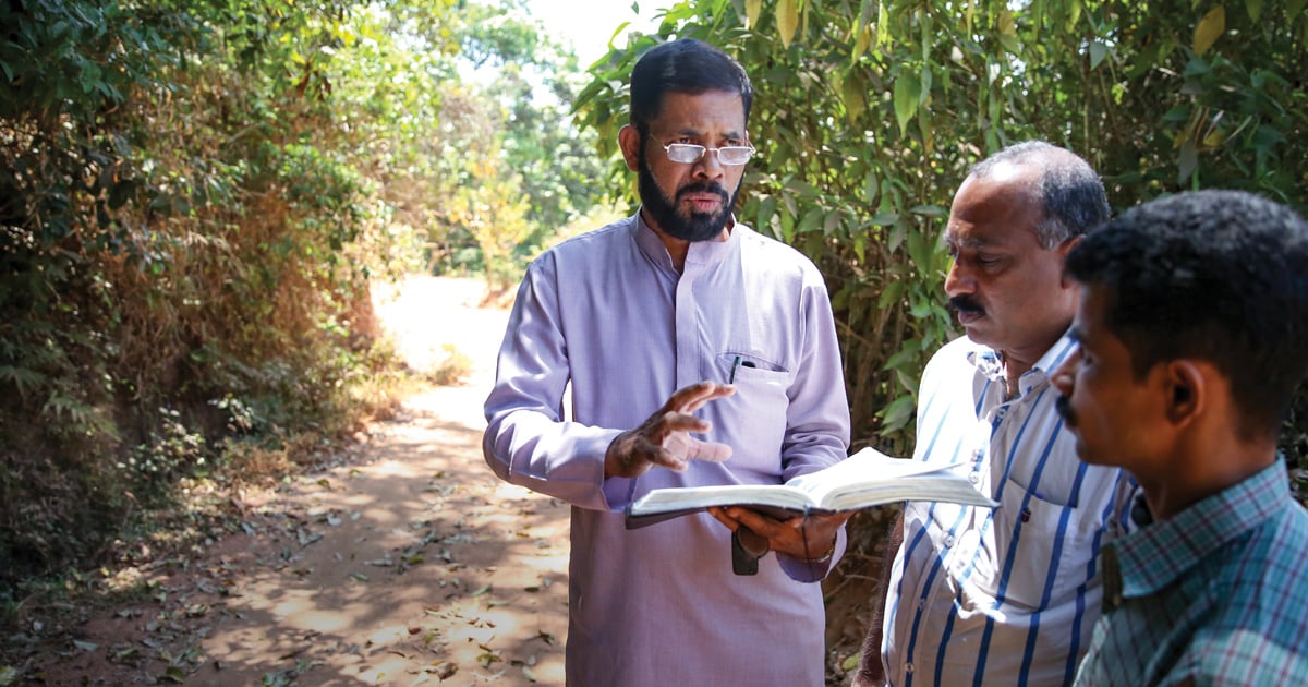 Three men are standing on a shaded pathway. One of the men has his Bible open and is speaking with the other two.