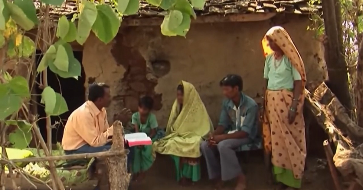 A man is sitting with his Bible open. He is speaking to a group of people.