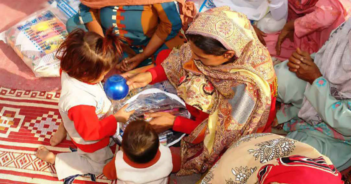 A woman and two children are looking through a small bag of goods. Other women are seated around them.