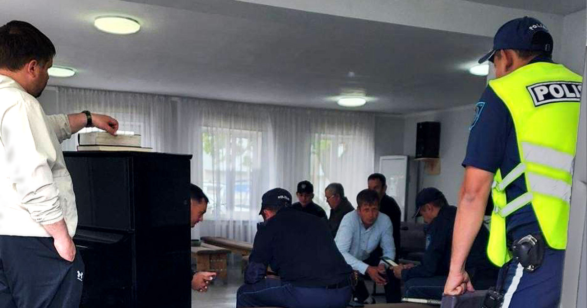 People are seated on benches. A police officer is standing and facing the group. People are seated on benches. A police officer is standing and facing the group.