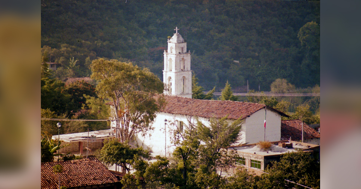 A white church building is surrounded by trees with a hillside in the background.