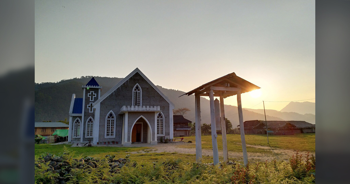 A small church building is in the foreground. The sun is seen just above the hilltops in the background.