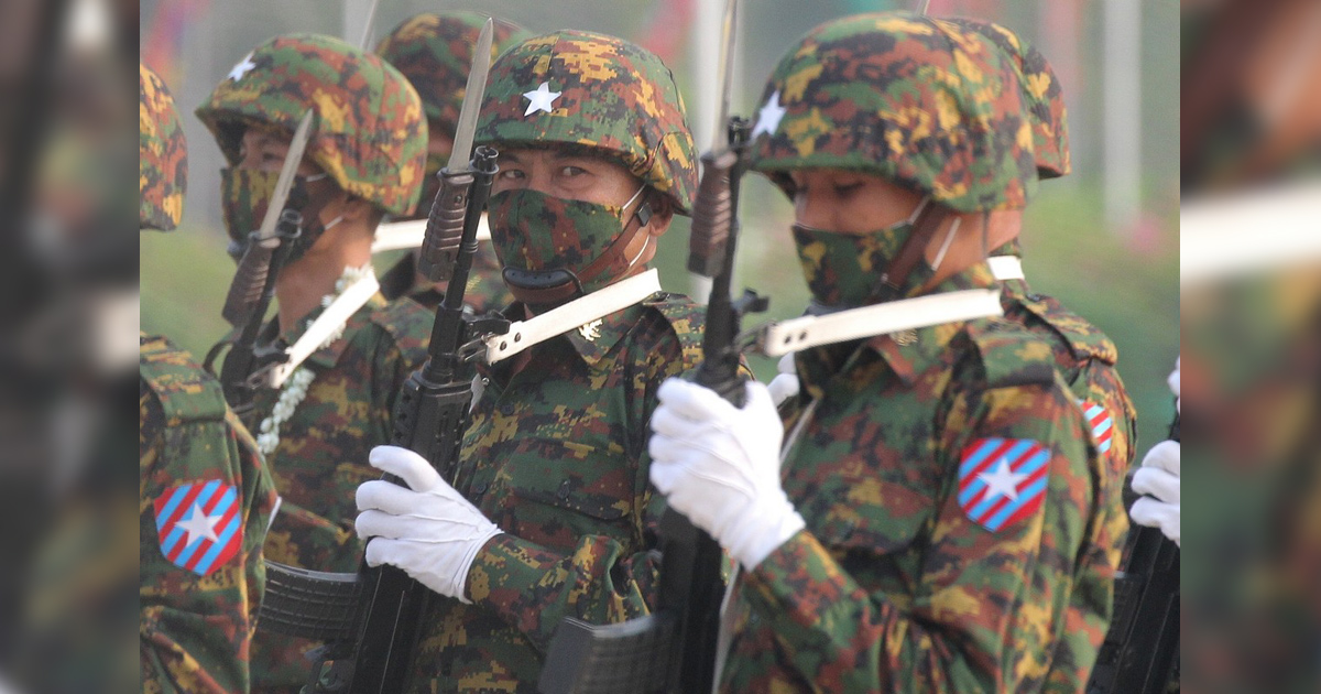 Photograph of Myanmar soldiers in camouflage uniforms and helmets during a military parade. White gloves, rifles, helmet markings, and red-and-blue shoulder insignia are visible.