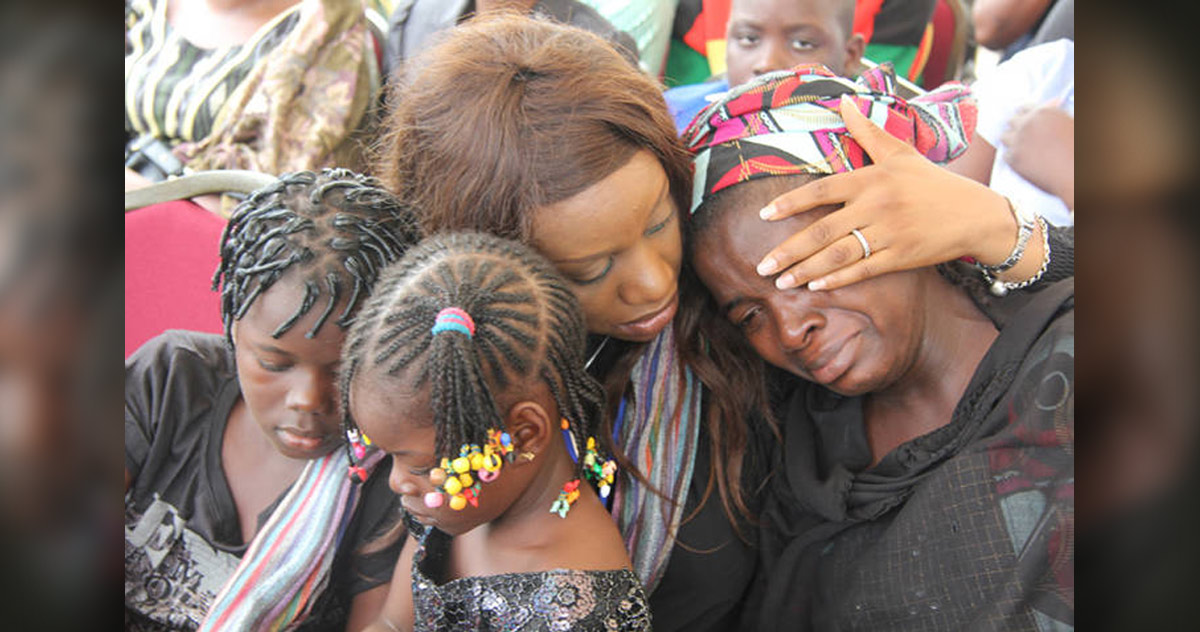 Photograph showing a woman comforting a tearful person wearing a colorful headscarf. Two children with braided hairstyles stand in the foreground, while adults in varied clothing fill background, conveying shared sorrow and support.