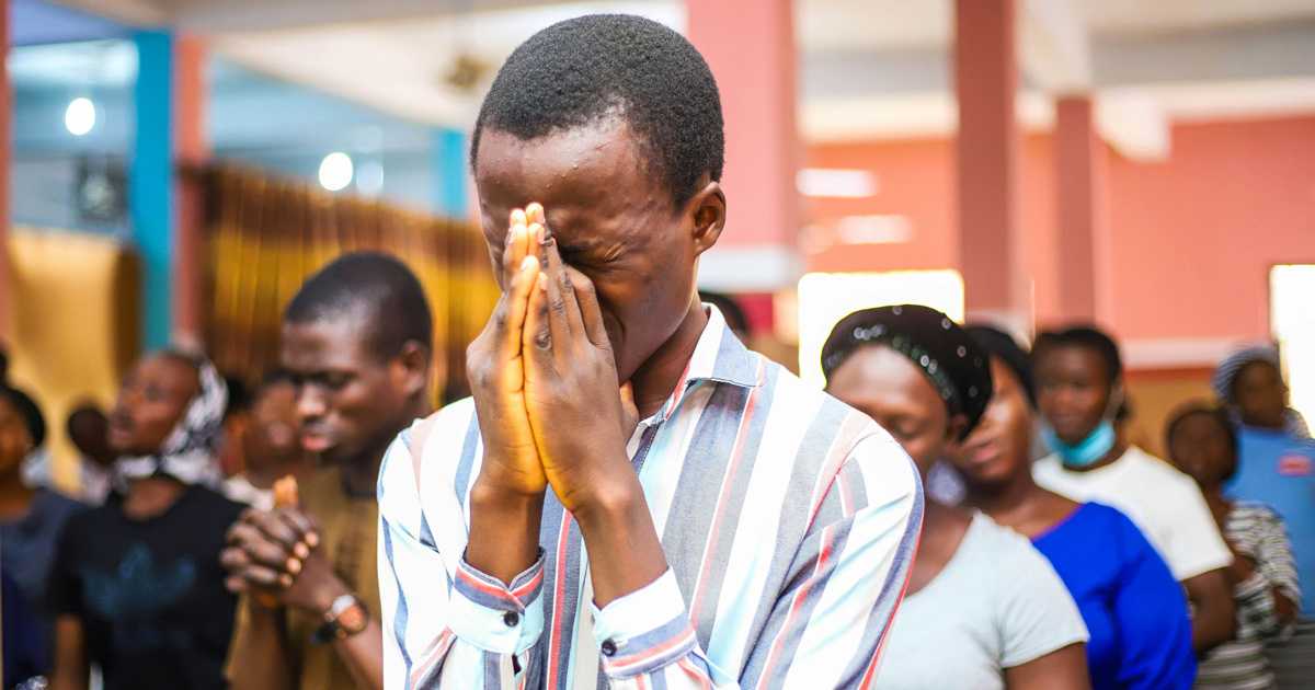 Several people are gathered together in prayer. The center of the image focuses on a man with his hands together and covering his face. He is focused on prayer.