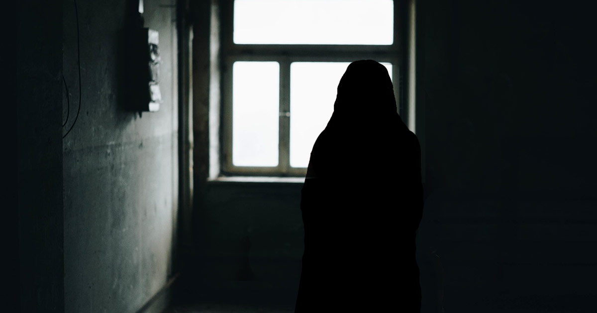 The silhouette of a woman in a head covering can be seen against a window. The room around her is very dark except for the daylight coming from the window. The silhouette of a woman in a head covering can be seen against a window. The room around her is very dark except for the daylight coming from the window.