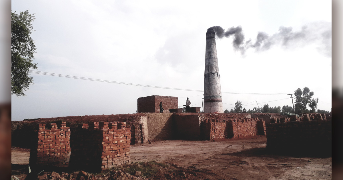A brick kiln has a very large, smoking chimney which is surrounded by brick buildings.