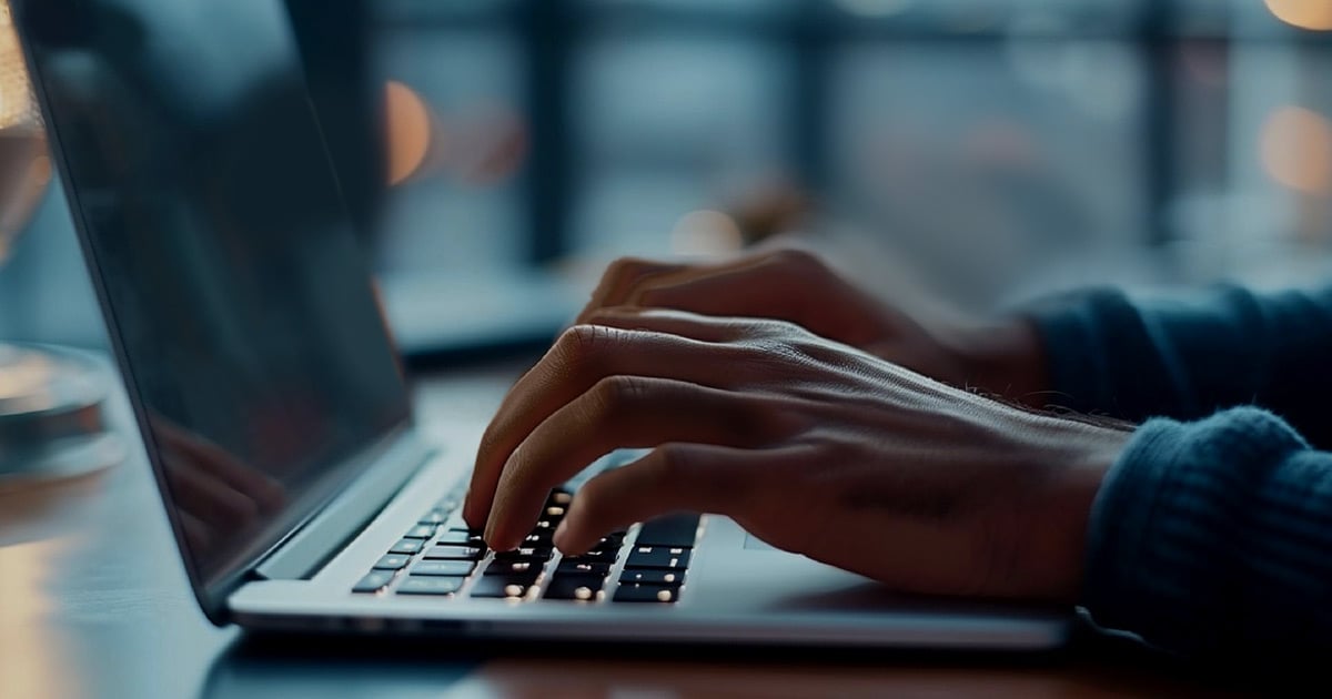 A man's hands are shown typing on a laptop in a dimly lit room.