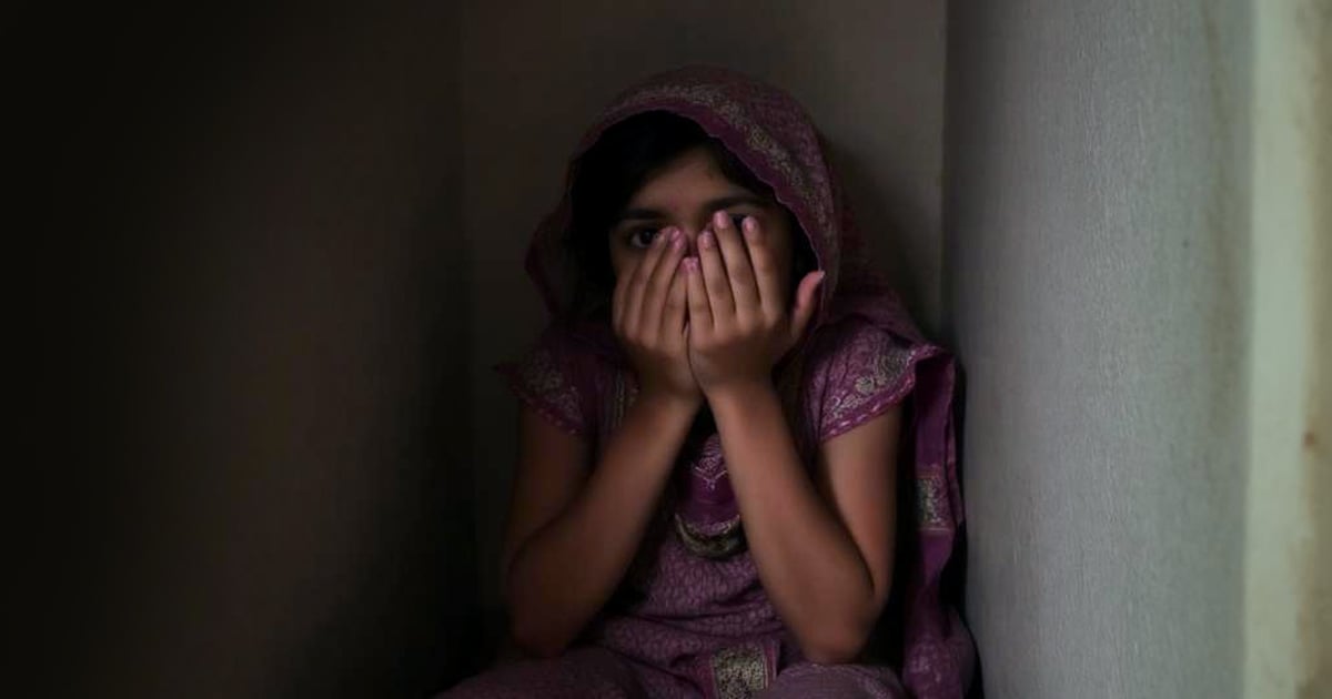 A teenaged girl is sitting in the corner of a dark room. She is covering most of her face with her hands.