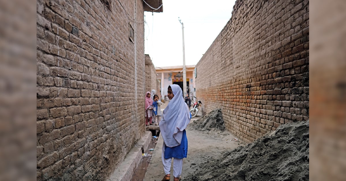 A girl is glancing back as she is walking between two brick buildings. There are more girls in the distance.