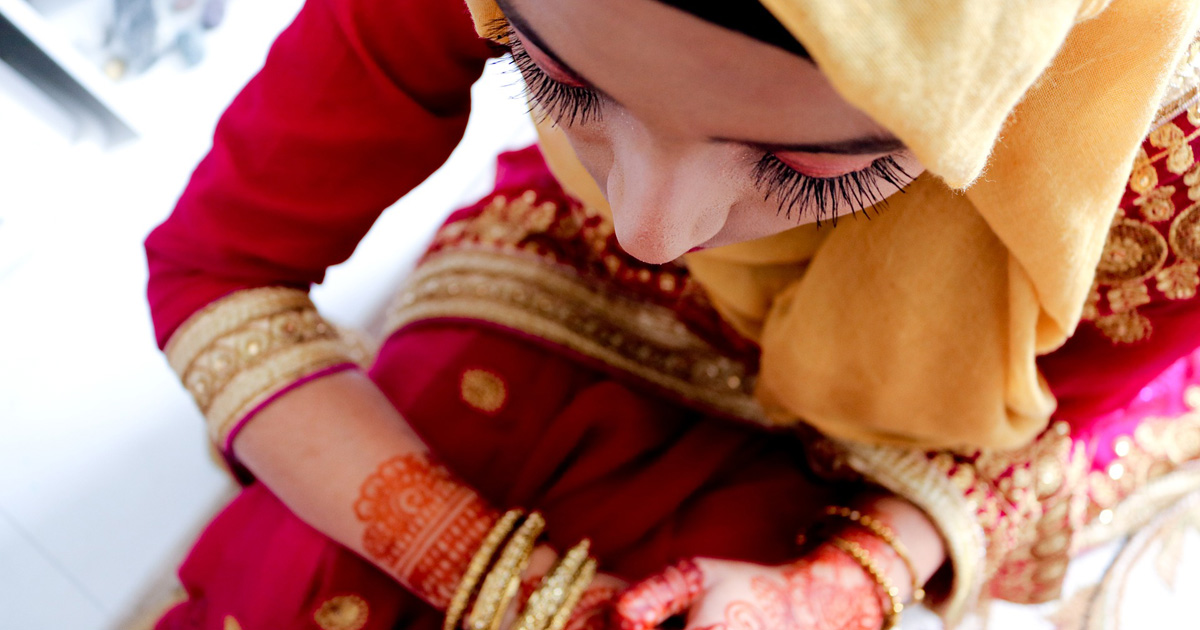 A Muslim bride sits with her head down. She is dressed in brightly coloured clothing and henna decorations cover her hands.