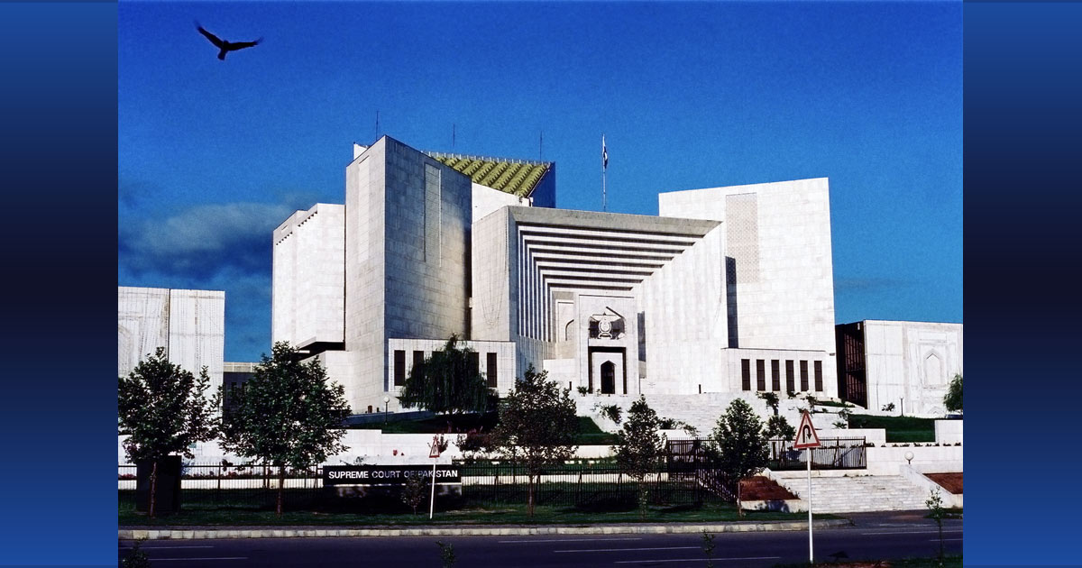 Photograph of a large white building featuring stepped geometric façades, deep rectangular recesses, and a central entrance. Blue sky, flag, trees, landscaped grounds, roadway, and flying bird frame the structure. Photograph of a large white building featuring stepped geometric façades, deep rectangular recesses, and a central entrance. Blue sky, flag, trees, landscaped grounds, roadway, and flying bird frame the structure.