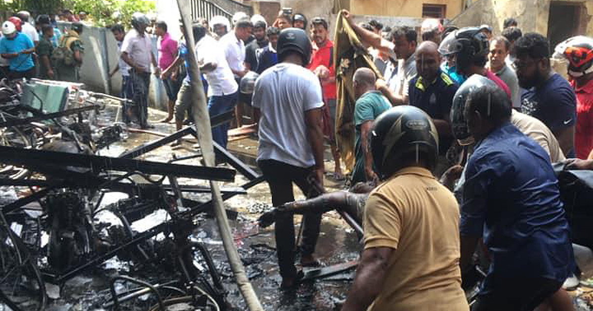 Photograph shows a crowded emergency scene with charred motorcycles, twisted metal, and debris after a fire. Bystanders and responders gather around the wreckage. Photograph shows a crowded emergency scene with charred motorcycles, twisted metal, and debris after a fire. Bystanders and responders gather around the wreckage.