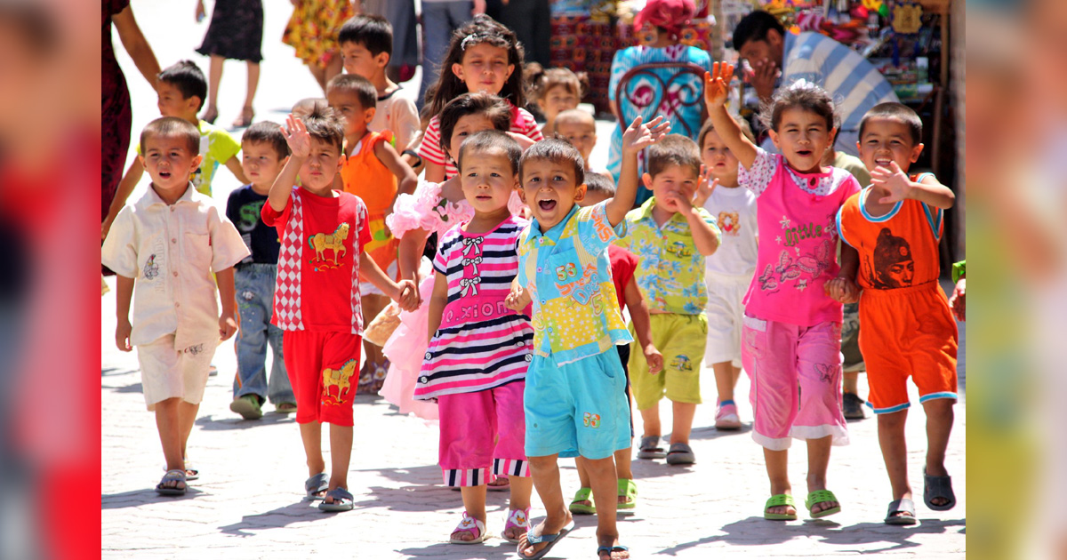 A group of happy children dressed in bright clothes greet the onlooker with waves and smiles.