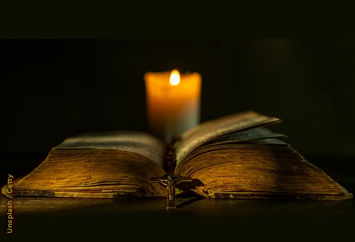 An open Bible is illuminated only by the light of a single candle behind it.