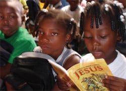 Children sit together. One girl is reading a book with the title, "Jesus."