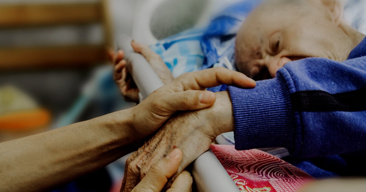 A frail, elderly man is reaching over a bed rail. Two caring hands are holding his.