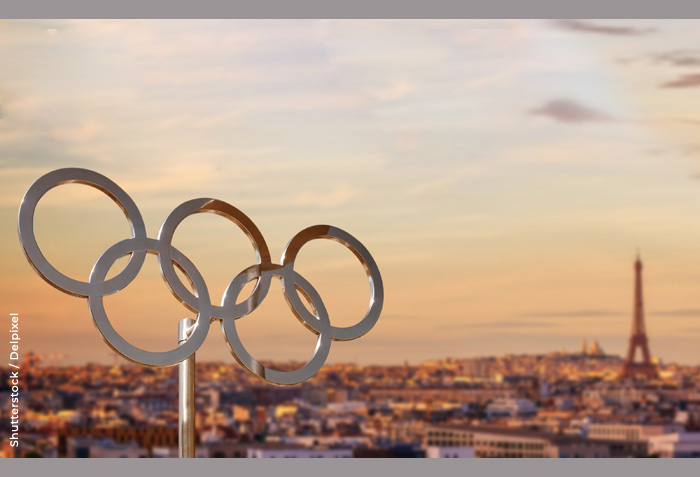 Metal Olympic rings are in the foreground. Paris is seen in the background.