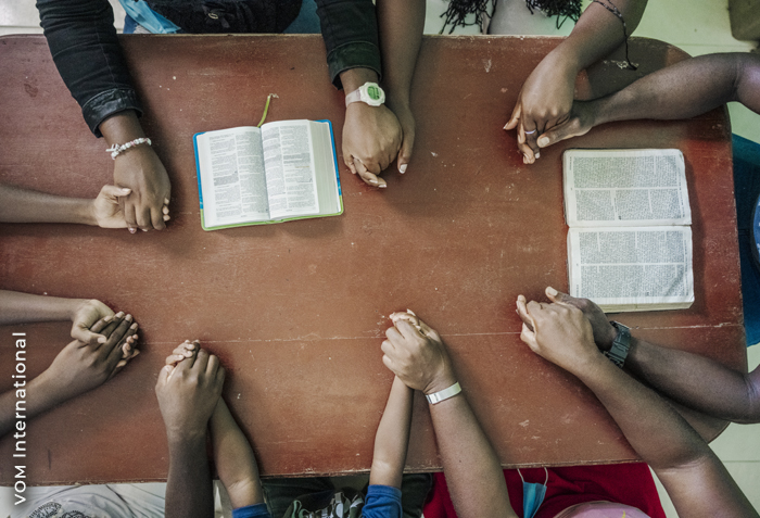 Seven people are sitting around a table holding hands. Bibles are open in front of two of them.