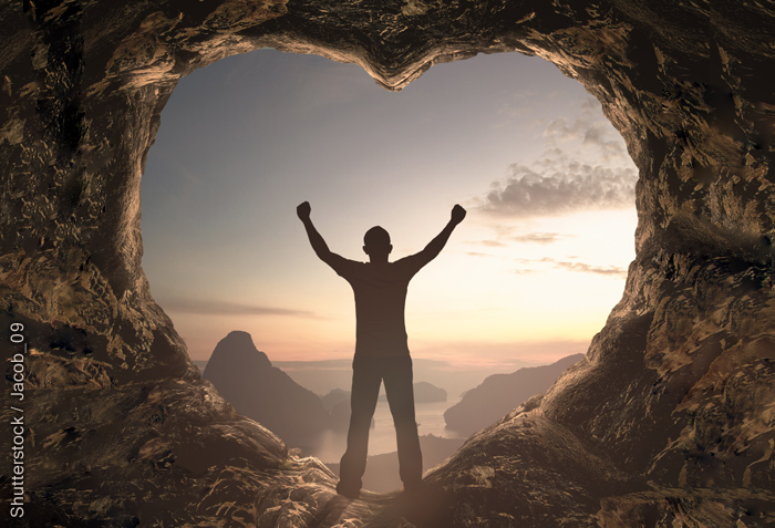 A man is standing in a heart-shaped cave opening with his arm raised. He faces a scene that is awash with the sun's rays on the horizon.