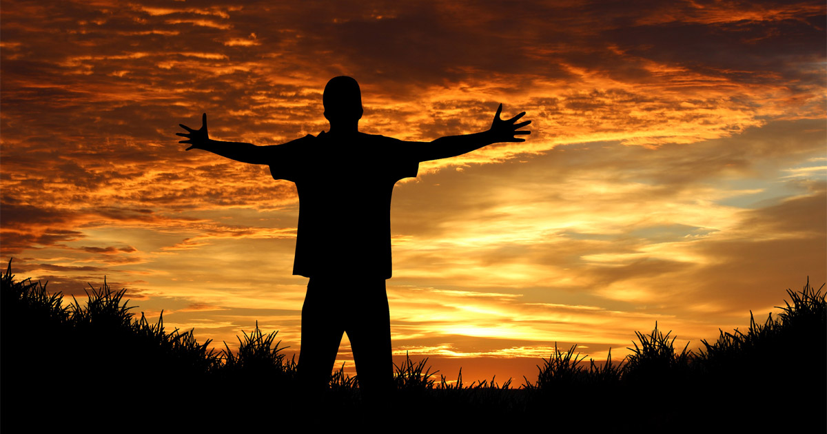 A man is standing with his arms raised. A sunset in the background creates a silhouette of him and the surrounding grass.