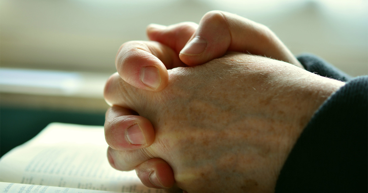 Hands are folded in prayer and resting on a Bible.