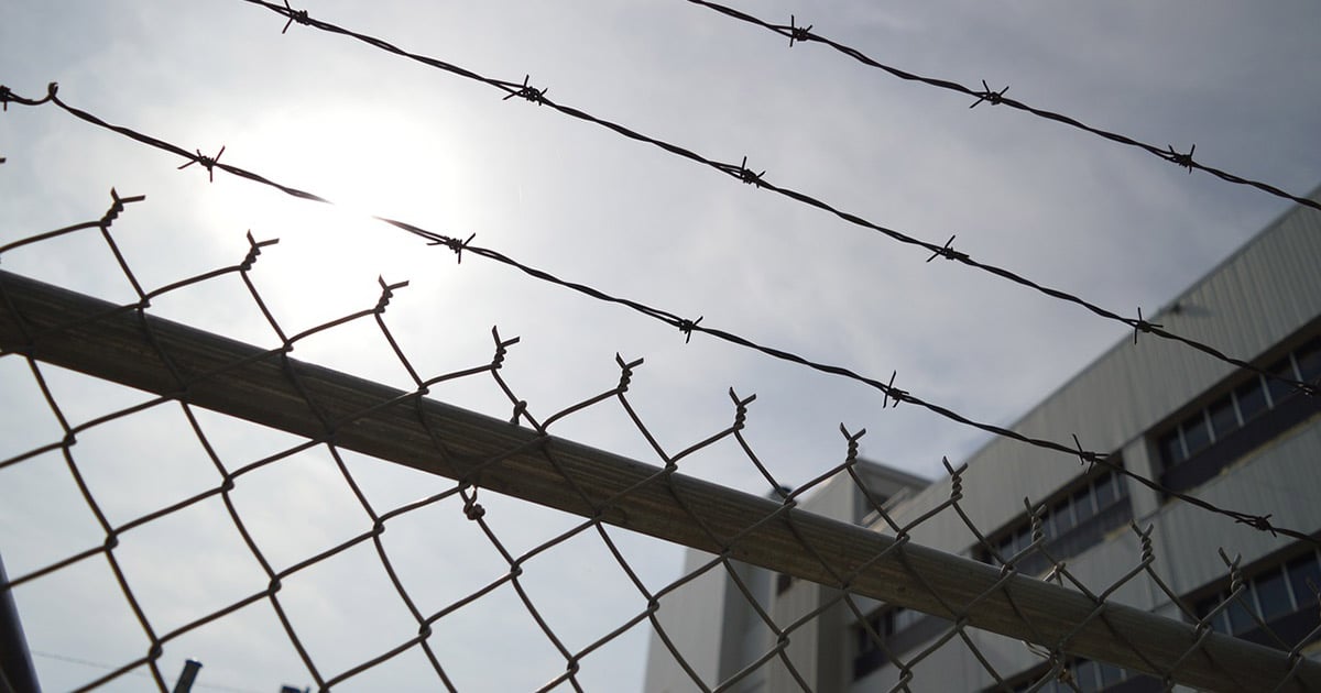 A building is behind a chain link fence with barbed wire security at the top.