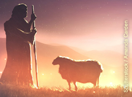 A silhouette of a shepherd and a sheep are standing on a hillside with strong light in the background.