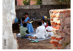 A Sunday School teacher is seated in front of children gathered on a piece of plastic laid out on the ground. They are in a building that has been largely destroyed.