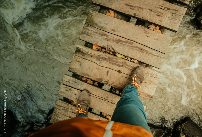 The feet of someone walking across a narrow foot bridge; water is swirling underneath.
